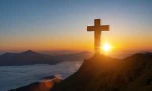 Schattenriss eines Gipfelkreuzes auf einem Berg vor einem Sonnenuntergang als Symbol für Bestattungsarten